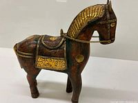 Side and angled view showing the wooden horse figurine with gold accents on the mane, saddle, and trim, including a gold chain around the neck.