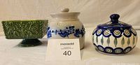 Lot of three ceramic and stoneware containers on display, including green footed bowl, white stoneware jar with blue flowers, and handmade jelly jar from Poland.