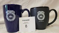 Two ceramic coffee mugs with Lower Merion Police Department logos, one dark blue and glossy, the other matte black, photographed together on a light cloth background
