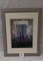 Framed photo of rustic stone wall with blue wooden doors and purple flowers in front