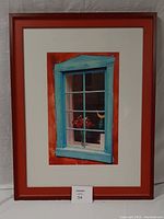 Framed photo showing aqua blue window and red-orange wall with visible flowers inside the window frame. The piece is vertically oriented and placed against a light wall.