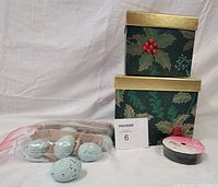 Photo showing two stacked decorative holiday gift boxes with holly and poinsettia motifs, a spool of black grosgrain ribbon, and a plastic bag holding six blue speckled decorative eggs.