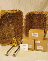 Two nesting baskets, two brass-colored Pottery Barn wall hooks, and four cardboard boxes with string lights on a beige fabric background.