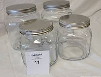 Four clear glass storage jars with silver-colored metal screw-on lids against a white background. Two jars are larger medium size, two are smaller size.