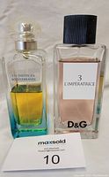 Photo of two fragrance bottles: Dolce & Gabbana L'Imperatrice partially used 3.3oz bottle and Hermes Un Jardin en Mediterranee half used 3.3oz bottle, with MaxSold bidding card.