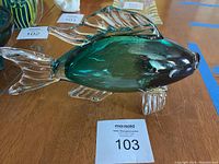 Green glass fish shown from left side with clear fins and tail, displayed on a wooden surface with auction tag 103.