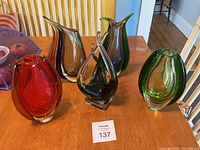Overview photo of five colored art glass pieces including four vases and one sculpture on wooden table