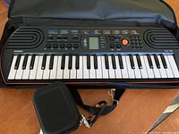 Casio SA-76 keyboard inside the open carry case showing 44 mini keys, control buttons, LCD display, and two speakers. The small black adapter case is next to the keyboard.