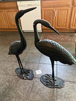Pair of black metal crane statues standing upright on a tiled floor with wooden kitchen cabinets behind.