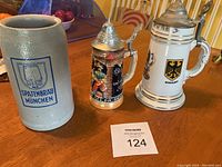 Photo showing the three steins on a wooden surface, including the large grey ceramic Spatenbräu stein, a small colorful stein with metal lid featuring a man in traditional attire, and a white ceramic stein with German eagle and metal lid.