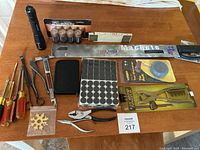 Photo showing a large machete, zoom flashlight, various screwdrivers, leather puncher, pliers, adhesive felt pads, a small zippered case, and multi-tool snowflake key on a wooden surface.