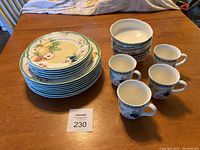 Stack of 8 dinner plates, 5 side plates, 4 bowls, and 4 mugs arranged on wooden table showing overall quantity and condition.