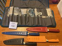 Top down view of cooking utensils laid out in a cloth roll with plastic packaging covering each utensil; four colorful knives with sheaths in front.