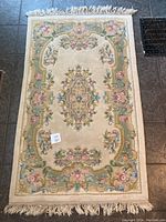 Full view of the rectangular wool carpet showing the floral and ornamental pastel design on ivory background with fringed edges, laid out flat on tiled floor.