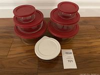 Two sets of Anchor glass storage bowls with red lids, plus one CorningWare glass dish with white lid, displayed on floor.