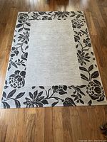 Top-down view of area rug laid flat on wood floor, showing light center with dark floral border.