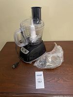 Black and Decker food processor with motor base, clear plastic bowl with handle and feed tube, and an additional plastic accessory wrapped in plastic on wooden surface.