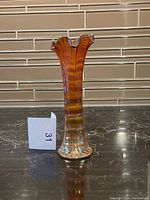 Photo of an amber ribbed glass vase with a ruffled top, placed on a dark marble surface in front of a tiled backsplash, with a small numbered card beside it.