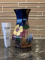 Front and side view of a glossy ceramic vase with hand-painted flowers in yellow, purple, brownish, and green on a dark background. The vase is approximately 8 inches tall with a flared top and slightly rounded body.