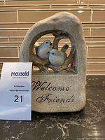 Stone decorative welcome ornament with two birds perched inside an arched frame and 'Welcome Friends' inscription, measuring about 5" W x 9" H.