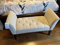 Beige fabric upholstered bench with tufted seat and curved armrests, standing on dark wooden legs, positioned on wood floor next to bed with floral bedding.
