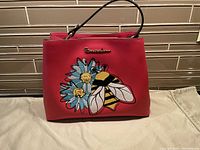 Front view of red Braccialini handbag featuring embroidered blue and white flowers and large bee with yellow and black stripes.