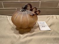 Glass pumpkin sculpture with amber textured body and curly clear glass stem with brown accents, shown against a tile backsplash.