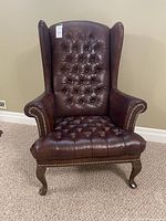 Front view of brown leather tufted wingback chair showing high back, rolled arms, nailhead trim, and Queen Anne legs