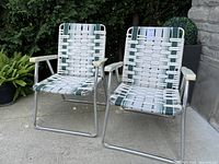 Two vintage lawn chairs shown side by side outdoors on concrete. Silver tubular metal frames with woven green and white plastic webbing seats and backs and white plastic armrests.