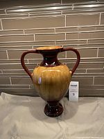 Photo of an ornamental ceramic vase with dual handles, set against a tiled background, showing front view and full shape.