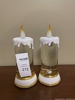 Pair of clear tall glass waterglobes shaped like candles, with white dripping wax effect on top, flame-shaped golden tinted tips, and gold accented white bases, measuring 11 inches tall.