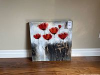 Photo of a square canvas painting with red poppies on a textured neutral background, placed against a beige wall on a wooden floor.