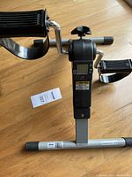 Side view showing the pedals with adjustable foot straps and the digital monitor mounted on a silver frame with black elements on a wooden floor.