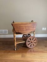 Side view of antique wooden tea cart folded, showing large wooden wheels and turned legs.