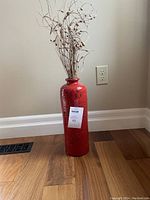 Red ceramic vase with dried flowers placed on wooden floor against beige wall.