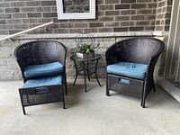 Full view of two dark brown wicker chairs with blue cushions, one with hidden pull-out foot stool extended, and round glass top wicker table between them.