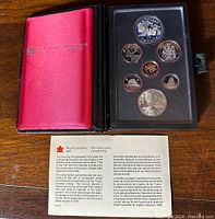 Opened red and black Royal Canadian Mint case showing seven coins, including a large silver dollar commemorative coin with locomotive design, in protective plastic slots.