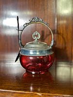 Front view of cranberry glass jam jar with silver plated lid, ornate handle, and spoon attached.