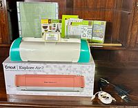 Cricut Expression 2 cutting machine in white and teal with accessories including cutting mats, cartridges, power and USB cords, all set on a Cricut Explore Air 2 box.