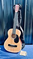 Full view of Denver acoustic guitar standing with gig bag and pack of strings on ground