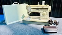 Full view of Singer 513 Stylist sewing machine with white hard case open behind it. Foot pedal and cords placed beside machine.