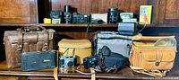 Wide shot showing a wooden shelf holding multiple vintage cameras, camera bags, lenses, and accessories arranged in two rows.