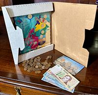 Vintage Canadian coin set displayed on colorful Canadian map in protective cardboard holder, along with loose coins and international banknotes on wooden surface.