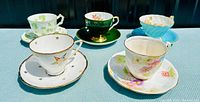 All six vintage fine bone china tea cups with matching saucers on a light blue cloth.
