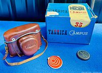 Photo of the Yashica Campus 35 camera case, original box, lens cap, and Japan Camera Industry Association tag, all displayed on blue felt.