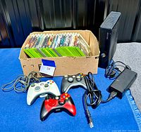 Xbox 360 console with power adapter, three controllers in front, and box of games behind