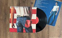 Vinyl LP record partly out of sleeve with cover featuring Bruce Springsteen's iconic Born In The USA imagery. Back inner sleeve with blue background and photo of Bruce Springsteen shown.