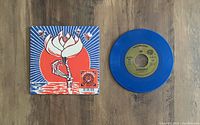 Blue 7-inch vinyl record next to full-color illustrated cover sleeve featuring dancing skeleton art and red sun design