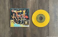 Yellow vinyl record with gold label and artwork cover of dancing skeletons with arrows in the background.