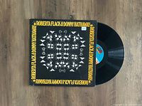 Black vinyl LP partially removed from its album cover showing front artwork with the artists' names and hand motifs.